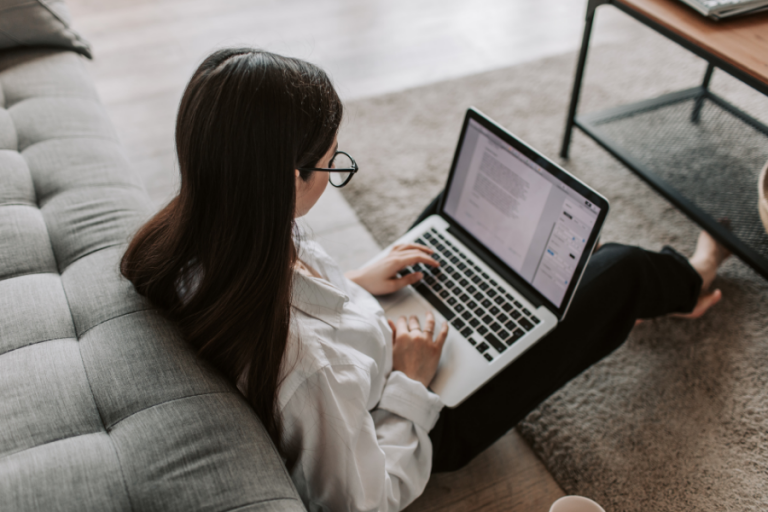 a woman working on her macbook sitting on her house floor, back against a sofa