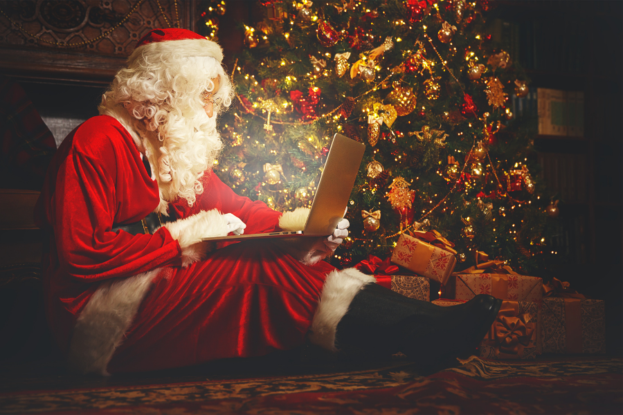 Santa claus using a laptop sat down on the floor by a Christmas tree